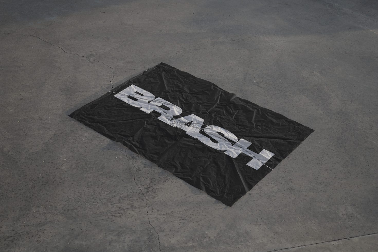 Black fabric with bold white text "BRASH" on a concrete surface for graphic design mockup, urban style presentation tool for designers.